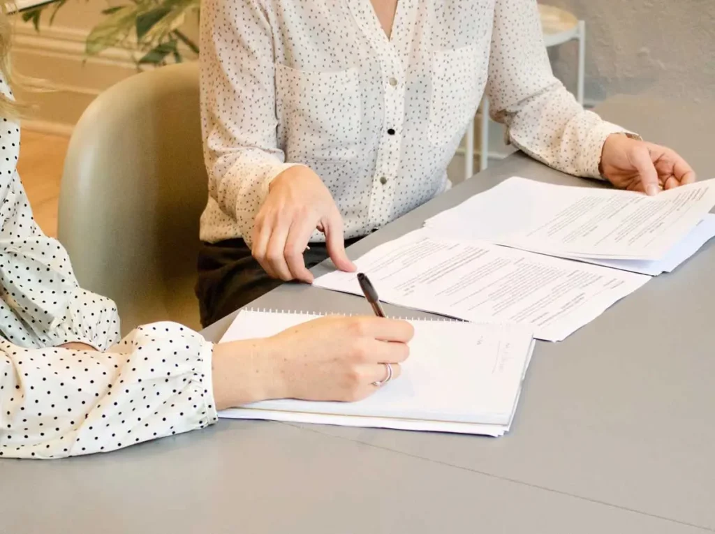 two women handling papers and writing in a notebook