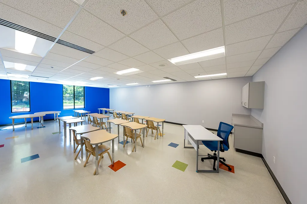 Classroom with desks and chairs. One bright blue wall with windows. Light gray walls, colorful tiled floor.