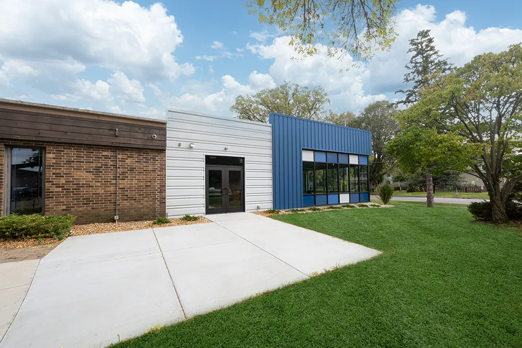 exterior view of building with brown bricks, blue and gray siding, double doors. Green grass and a sidewalk