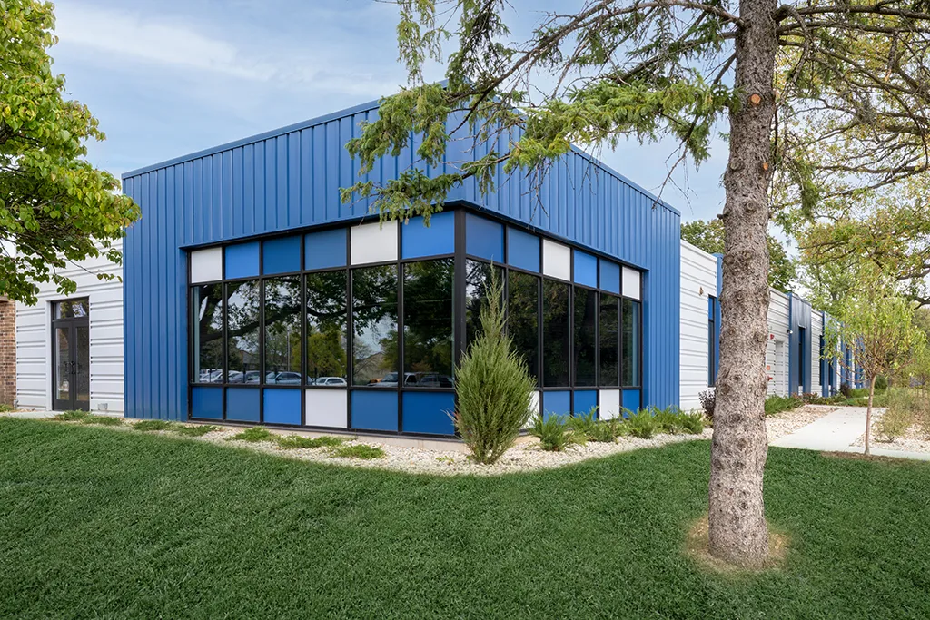 Exterior view of corner of building with blue and gray siding, big windows, green grass and a tree