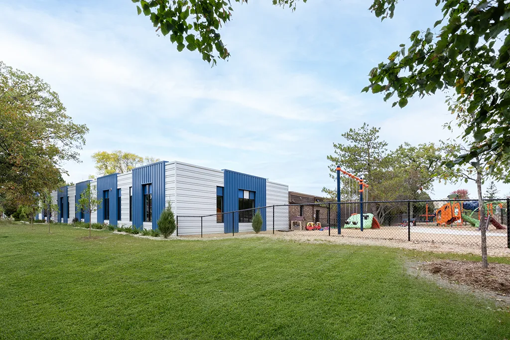 building with blue and gray siding, large windows. Green grass, a fence and colorful playground equipment