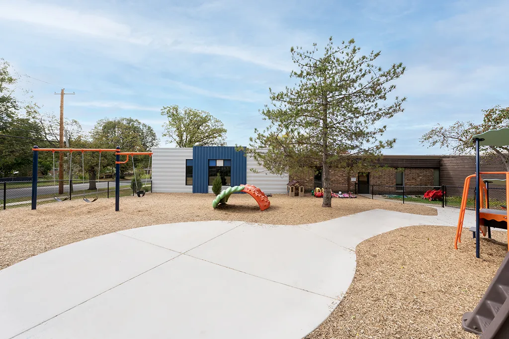 building with brown bricks, blue and gray siding. Playground with walkways, chipped wood, and trees