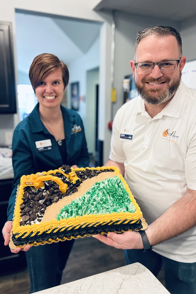 A woman and a man holding a sheet cake decorated to look like a construction site with two yellow trucks