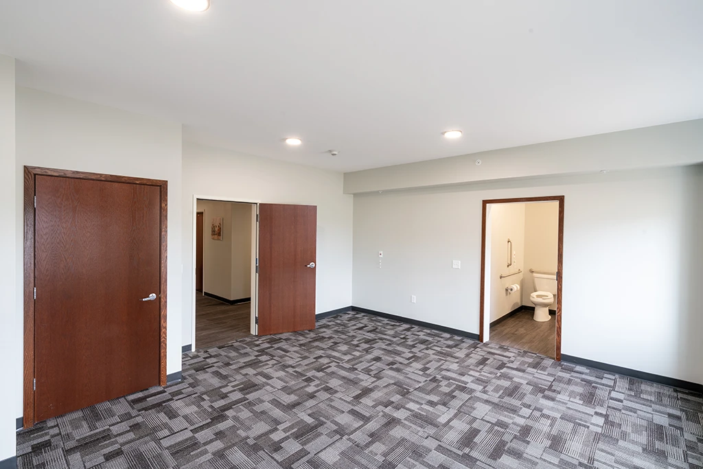 Large room with gray patterned carpet, brown doors, and a bathroom with white toilet