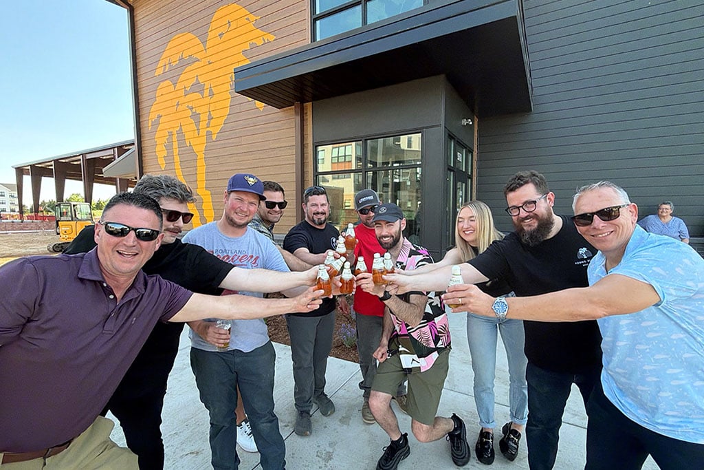 Group of people outside building with brown siding and orange palm trees on the side clink bottles of beer