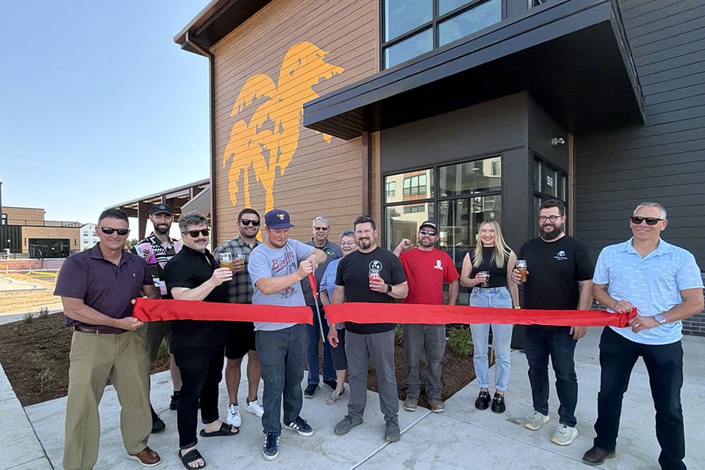 Group of people from Young Blood Beer Co and Supreme Structures cutting a large red ribbon in front of a brewery with brown siding and large orange palm trees on the side